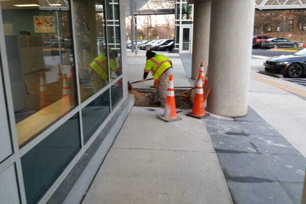A worker fixing part of a sidewalk outside of a building and parking lot.