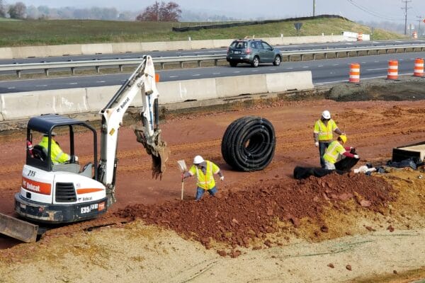 Workers on a jobsite with materials and machinery working on utilities in the middle of the highway with cars passing in the background.
